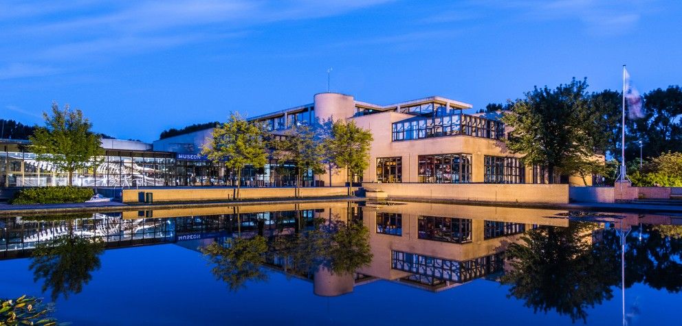 Het gebouw van Museon-Omniversum in de avond, met vooraan een groot wateroppervlakte waarin het museum wordt weerspiegeld.