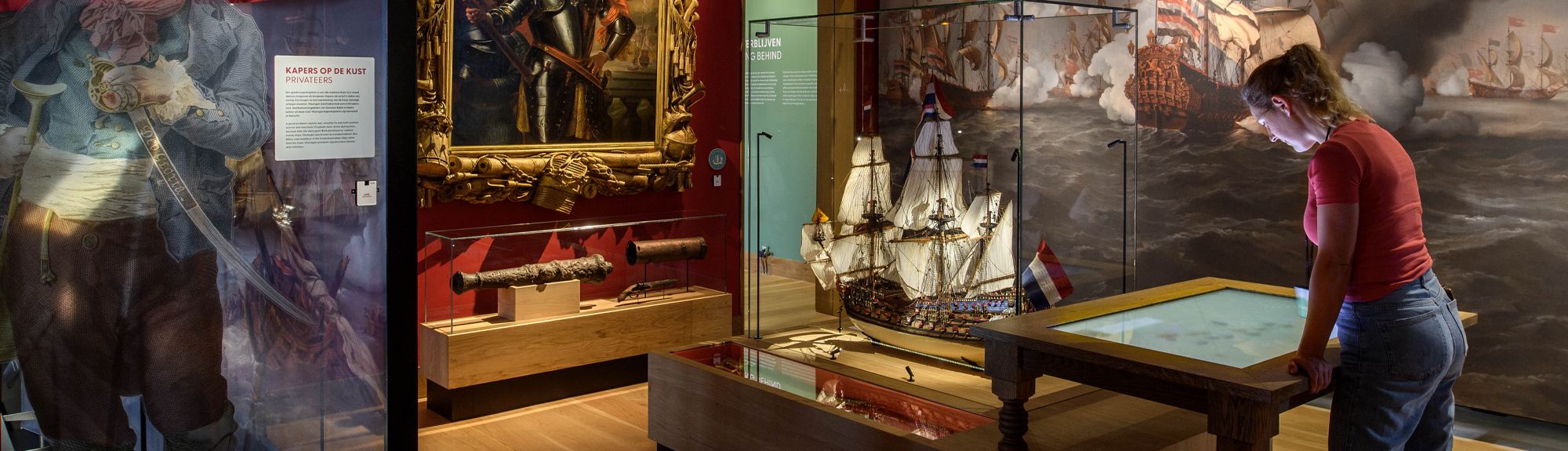 Een vrouw bekijkt een beeldscherm in een tentoonstelling over de zeevaart in Maritiem Muzeeum Zeeland. De ruimte is gevuld met historische stukken en een replica van een 17e-eeuws schip. Met de VIP-KAART bezoekt u dit museum gratis.