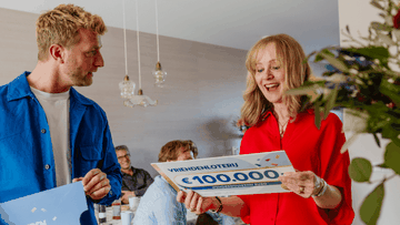 Een vrouw leest enthousiast een cheque van €100.000 van de VriendenLoterij, terwijl een man naast haar staat en anderen op de achtergrond zitten.
