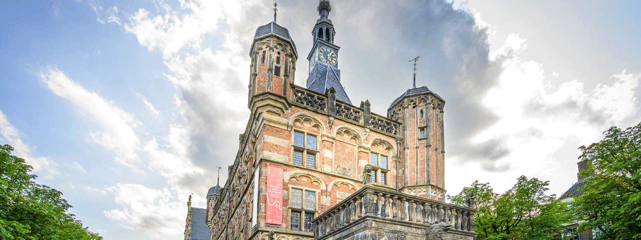 Een historische waaggebouw midden op een marktplein. Aan het bakstenen gebouw hangt een rode banner met Hanzestad Deventer. Ontvang gratis toegang tot Museum De Waag met de VIP-KAART