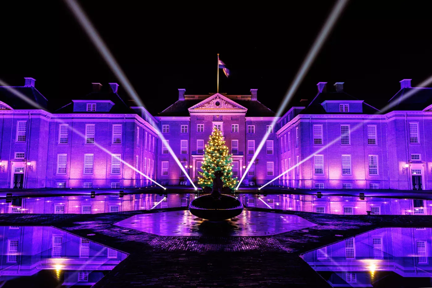 Een sfeervolle foto van Paleis Het Loo in de nacht. Het statige paleis wordt verlicht door paarsblauwe lichten. In het midden staat een grote kerstboom. U kunt dit bijzondere museum gratis bezoeken met de VIP-KAART.