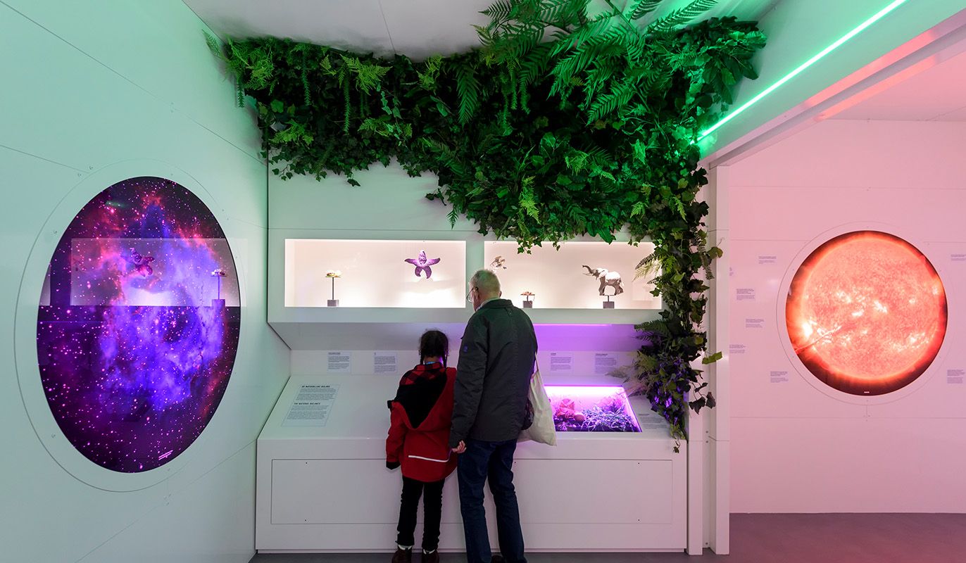 Twee bezoekers bekijken een interactieve tentoonstelling in Oyfo Techniekmuseum in Hengelo, met kleurrijke verlichting, ruimtebeelden en kunstobjecten onder een groen bladerdak. Gratis toegang met de VriendenLoterij VIP-KAART.
