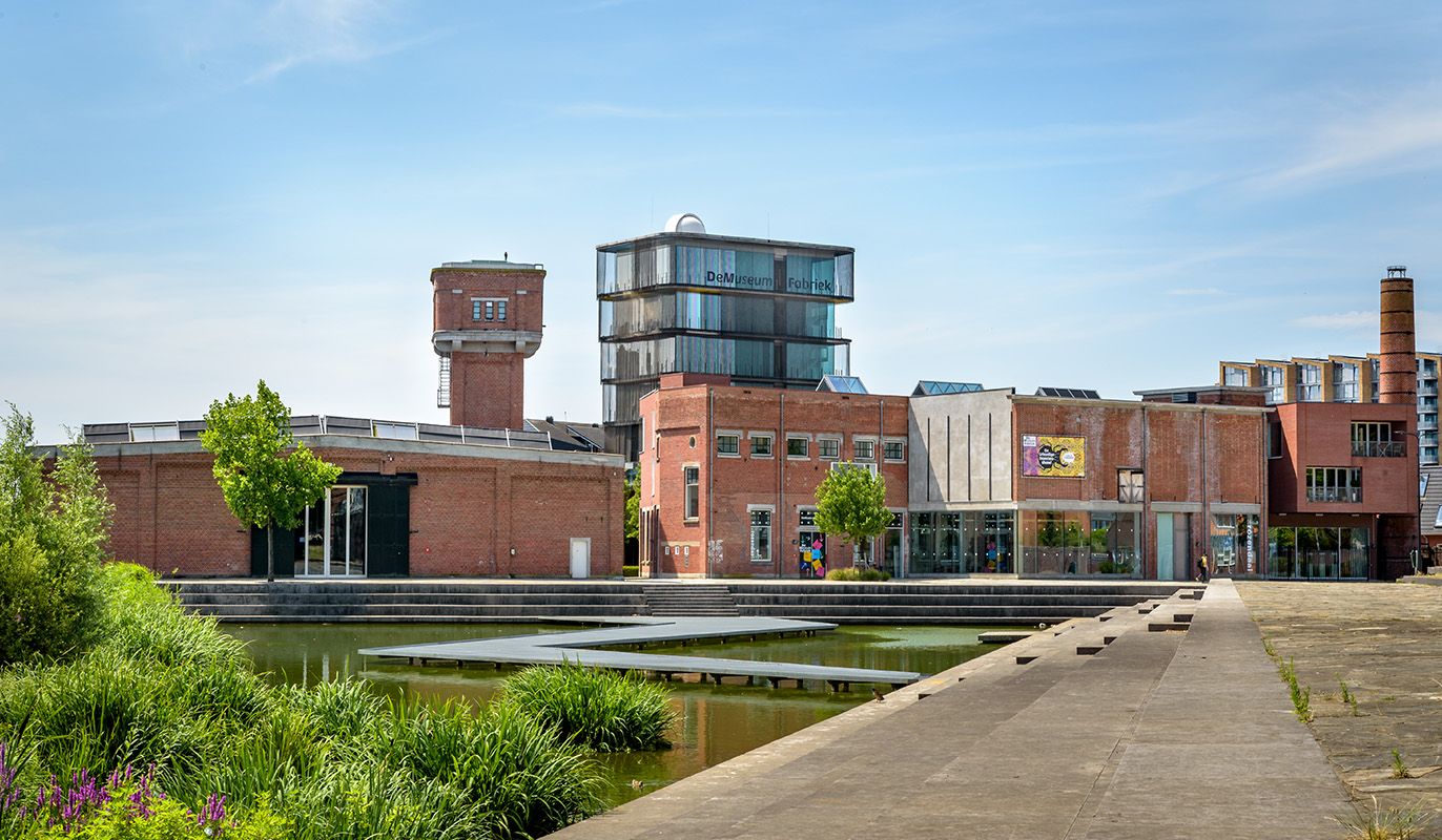 Exterieur van De Museumfabriek in Enschede, een creatief museum in een oude textielfabriek met moderne glazen uitbouw en een vijver op de voorgrond. Gratis te bezoeken met de VriendenLoterij VIP-KAART.
